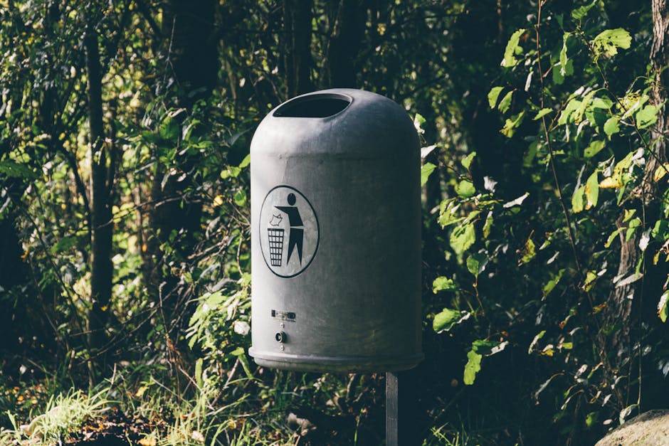 A cylindrical metal rubbish bin with a rounded top and a large oval opening at the top for waste disposal, mounted on a metal post in an outdoor environment amid dense green foliage and shrubbery. The surface of the bin appears weathered with a matte, metallic finish, and there is a black and white icon on the front depicting a person throwing trash into a bin, emphasizing proper waste disposal. The surrounding area includes a mix of leafy plants with various shades of green, and the lighting suggests natural daylight, casting soft shadows and highlighting the contrast between the bin's metallic surface and the lush greenery. This scene illustrates a location suitable for independent or private waste collection, typical of community or park settings where waste management services like those of Waste Collection Chiswick are relevant to maintaining cleanliness and environmental standards.