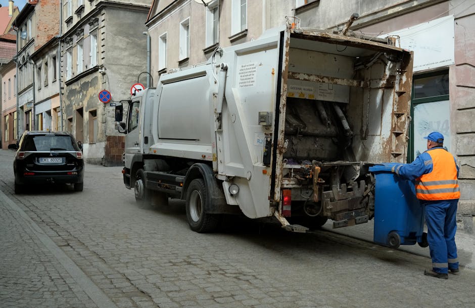 A street scene featuring a white waste collection truck with its rear compactor open, positioned on a cobblestone road in front of a row of multi-story buildings with aged facades. The truck's metal surface shows signs of rust and dirt, particularly around the rear loading area, which is designed for collecting bulk rubbish and debris. To the right, a worker dressed in a high-visibility orange vest with blue trousers and a matching blue cap is actively placing a standard blue plastic bin into the truck's hopper, suggesting a routine private waste collection operation. The background includes parked cars, including a black vehicle on the left side, and signage indicating parking restrictions mounted on the buildings. The overall environment appears urban, with natural light illuminating the scene, emphasizing the textures of the cobblestones, the truck’s metallic surfaces, and the worker’s clothing, all contributing to a professional waste management context associated with independent rubbish removal services like those offered by Waste Collection Chiswick.