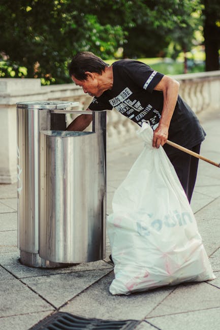 A photograph taken from the perspective of a person standing on a grassy area beside a paved path, showing a collection of scattered rubbish on the ground. The waste includes a crumpled brown paper bag, various pieces of food packaging such as a white plastic container, a red and white cardboard drink cup with a plastic lid, and a crumpled paper napkin. The person's white sneakers and lower legs are visible at the bottom of the image, indicating they are standing close to the debris. The surrounding environment consists of green grass, and the scene suggests a case of littering in an outdoor area near a pathway, highlighting the importance of proper waste disposal and waste management services like those provided by Waste Collection Chiswick.