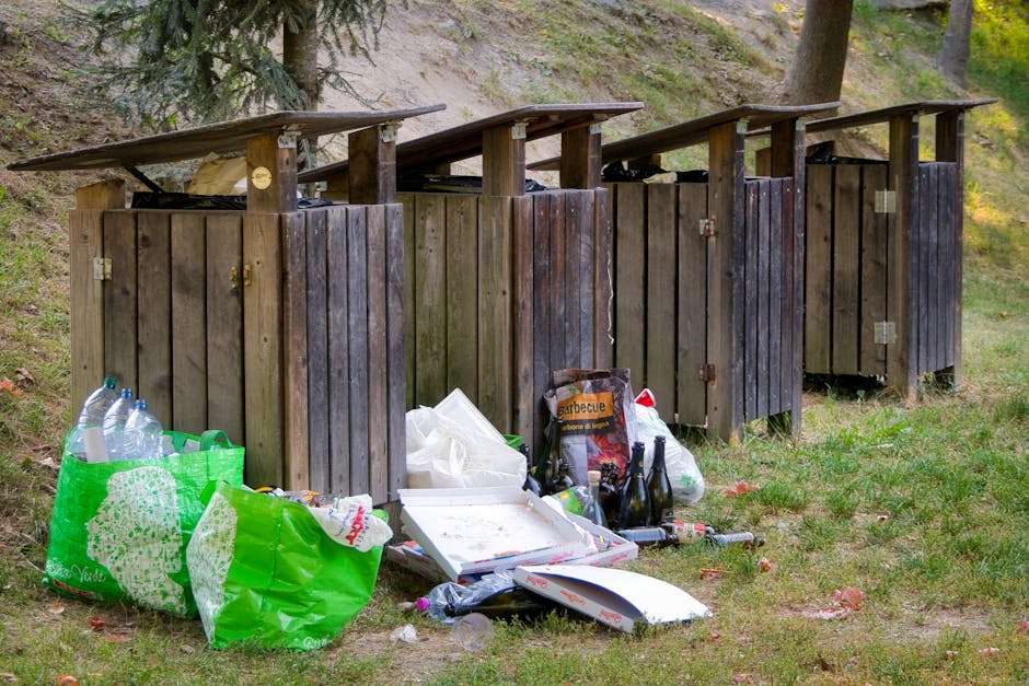 Four wooden outdoor rubbish bins with vertical slats and sloped roofs are positioned side by side on a grassy area next to a sloped, wooded hillside. The bins are dark brown, weathered, and appear to be used for waste collection. In front of the bins, various discarded items are strewn on the ground, including a large green plastic shopping bag filled with empty plastic bottles, a white cardboard pizza box with remnants of food, a black bag labeled 'barbecue' containing charcoal or grill supplies, and several glass bottles. There are additional loose waste items such as plastic wrappers and a small discarded object resembling a bottle opener. The scene suggests a private or informal rubbish disposal area, not a scheduled municipal collection point. The background features tall trees and patches of sunlight filtering through the foliage, contributing to an outdoor, natural environment typical of a residential or semi-rural setting. The image illustrates concerns around onsite waste management and unauthorized dumping that Waste Collection Chiswick may assist with through private rubbish removal services.