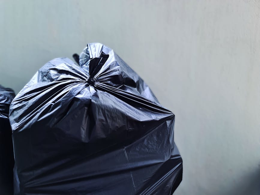 A collection of overflowing rubbish bins and scattered waste bags are placed on a paved sidewalk in front of a commercial building. The grey mixed paper and card bin lid is open, revealing paper, cardboard, and packaging materials protruding from inside. To its right, a black bin with a partially visible label holds additional waste, with black plastic bags piled nearby. A red bin beside it is also filled with waste, while some cardboard boxes and loose trash, including bottles, plastic containers, and paper, are strewn across the ground. Behind the waste collection point, a metal railing separates the area from a parked silver hatchback car on a designated parking space. The background features storefronts with yellow signage, and the upper level of the building is under construction, covered in scaffolding and blue protective sheeting, indicating ongoing development or maintenance work. The overall scene depicts a typical urban rubbish accumulation, illustrating the necessity of regular collection and waste management services provided by Waste Collection Chiswick, aligning with alternative disposal methods outside standard council pickups.